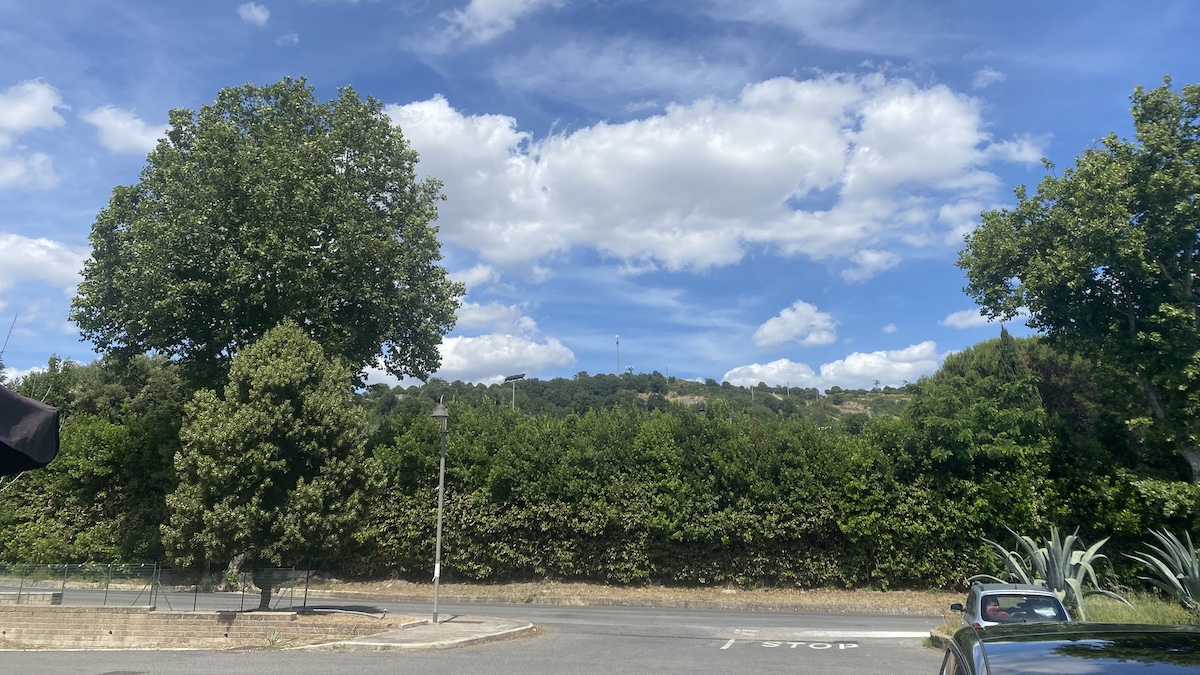 Summit of Monte Razzano. Car park exit with two large trees on either side, trees in the foreground and rising up to a summit in the distance on which is a mobile phone mast