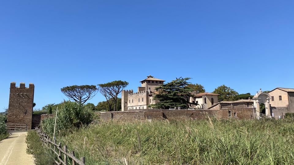 A large complex holding a crenellated church with an imposing marble gateway and a big cedar of lebanon. A small crenellated tower stands on the corner at the left, in front of the cycleway. 