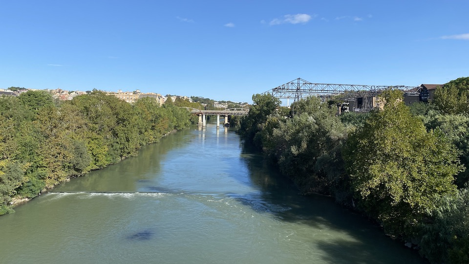 Looking down on the Tiber river. In the river there is a small weir with white water tumbling over it. The river banks are green, and there are the outline of an older bridge and the industrial remains of the old gasometer and unloading conveyor.