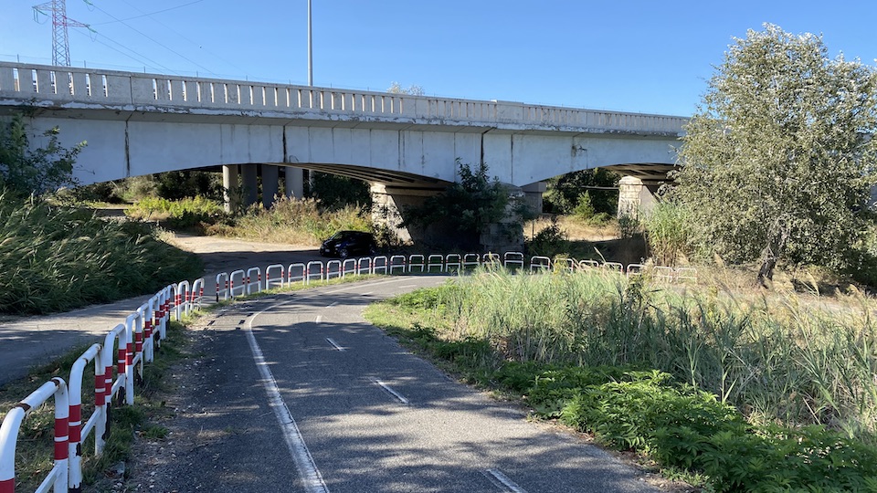 A concrete road bridge above the river. In the foreground, red and white rails line the bike path which ends just below