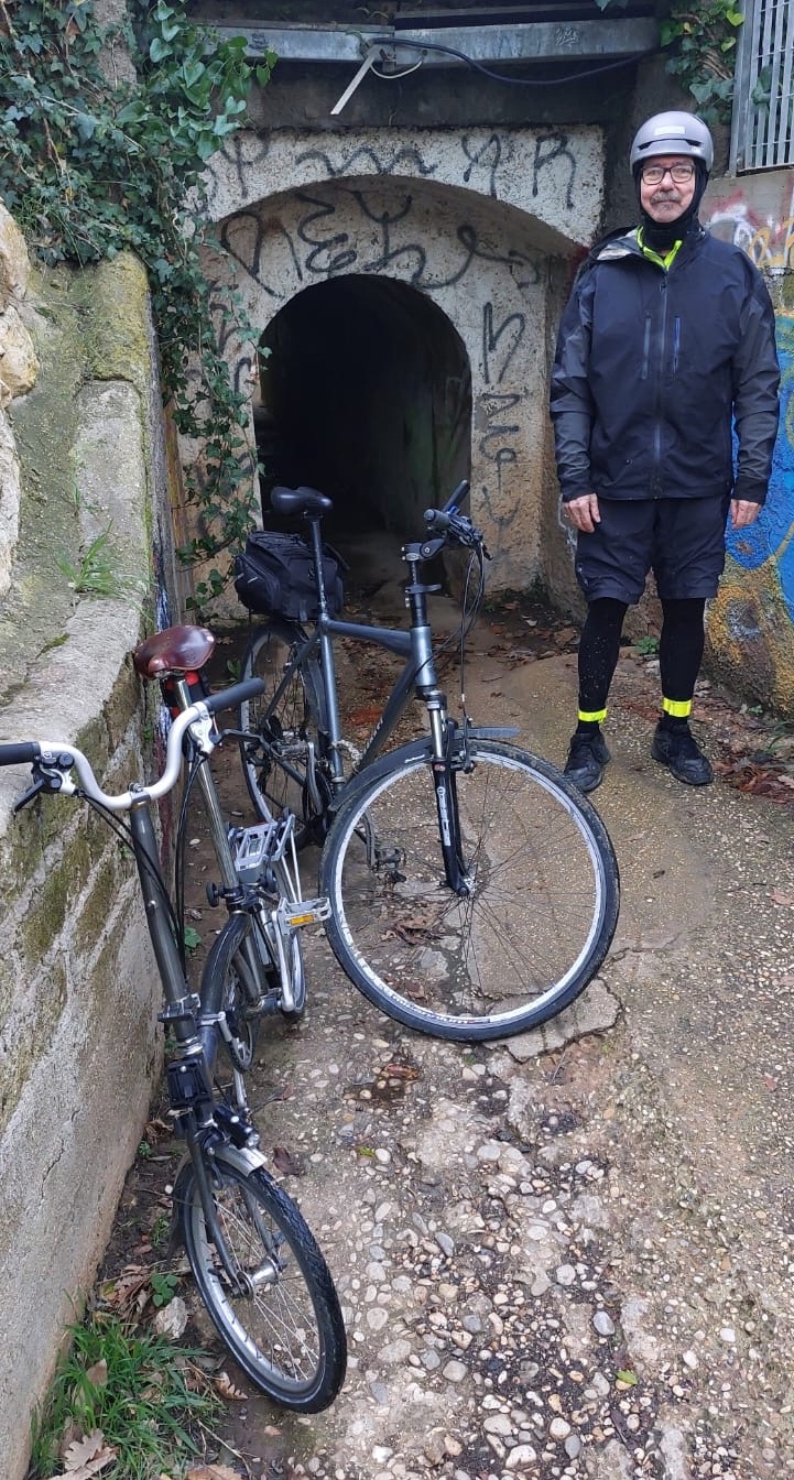 A cyclist, dripping wet, dressed in black with fluorescent yellow highlights, standing in front of a small grafittied tunnel about half his weight. In front of him are two bicycles, one of which is a Brompton