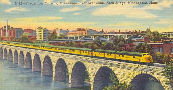 Old postcard of a Streamliner train, locomotive and cars are yellow with green roof, on the Stone Arch Bridge in Minneapolis, with the huge flour mills in the background