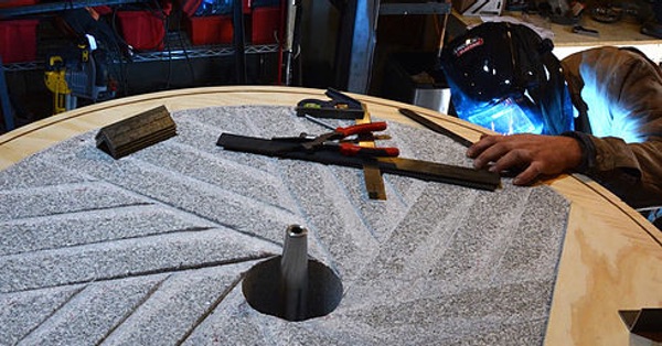 A New American Stone Mill under construction. In the foreground, the gray millstone incised with the grooves that will shear and then grind the wheat as it rotates. Behind, a man in a welding mask is working on the metal frame, his face lit by the intense blue arc of the welder. Sonme tools rest on top of the stone.