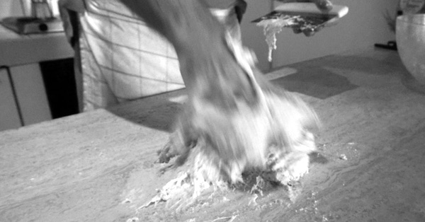 Monochrome photo of a person kneading bread dough on a wooden countertop. The hand and dough are in motion and blurred.